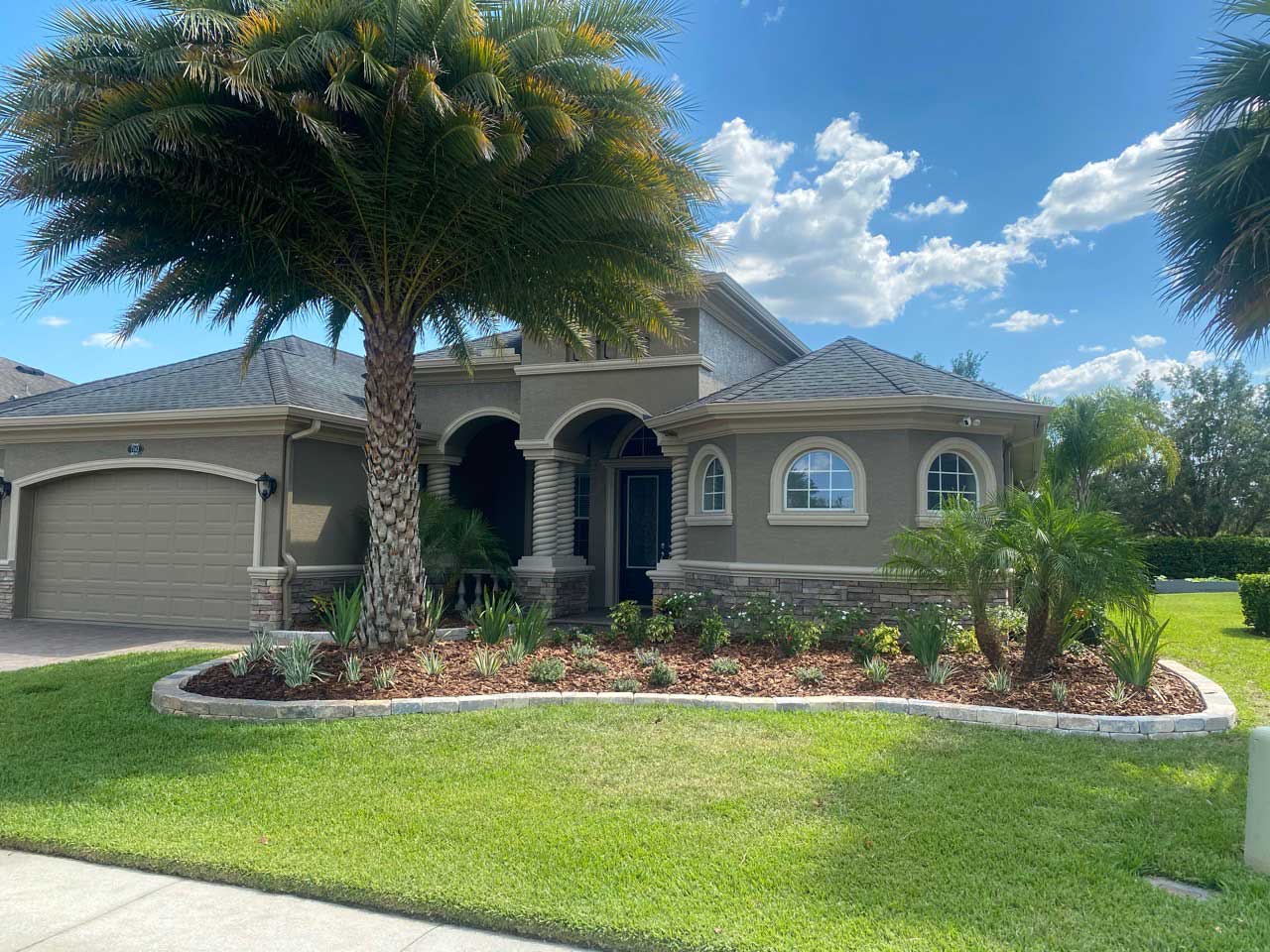 A beige, single-story house with arched windows and entryway features expert landscape design in the front yard, showcasing palm trees, lush plants, and a two-car garage under a sunny blue sky with scattered clouds.