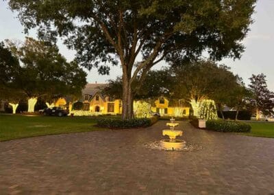 A large house features professional Christmas light installation, with white holiday lights on trees and around windows. The brick-paved driveway leads to a small, illuminated fountain surrounded by green lawns and tall trees.